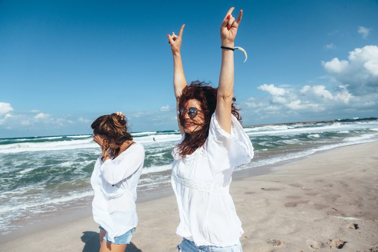 Δύο φίλες περπατούν δίπλα στη θάλασσα με ψηλά τα χέρια χαρούμενες