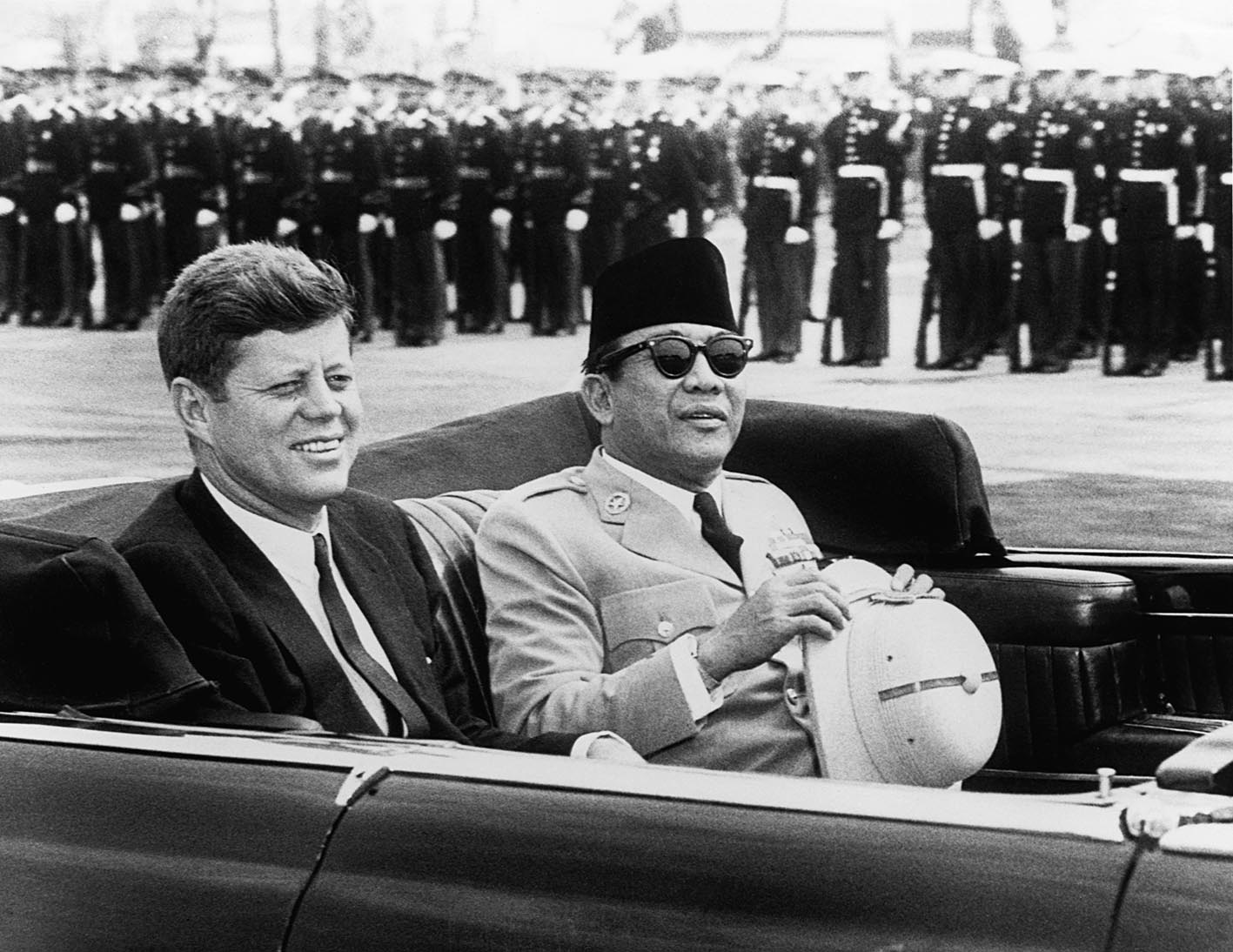 Ο πρόεδρος Kennedy και ο πρόεδρος της Ινδονησίας Sukarno σε ένα ανοιχτό αυτοκίνητο προσπερνώντας μια σειρά στρατιωτών.