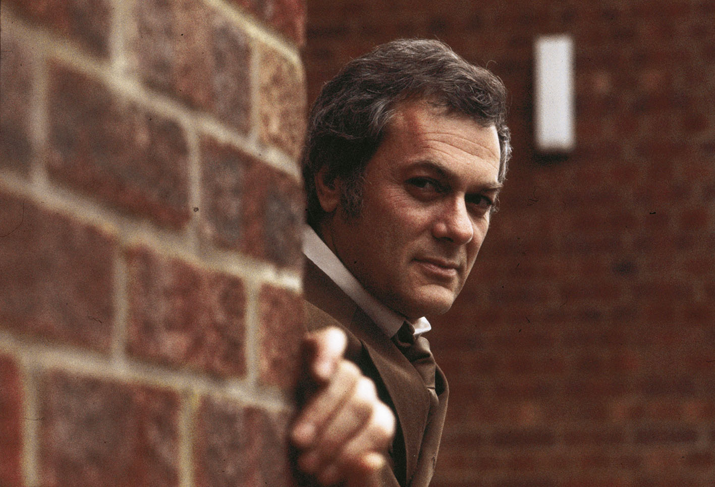 1970: Ο Αμερικανός ηθοποιός Tony Curtis κοιτάζει πίσω από έναν τοίχο κατά τη διάρκεια επίσκεψής του στο Λονδίνο.