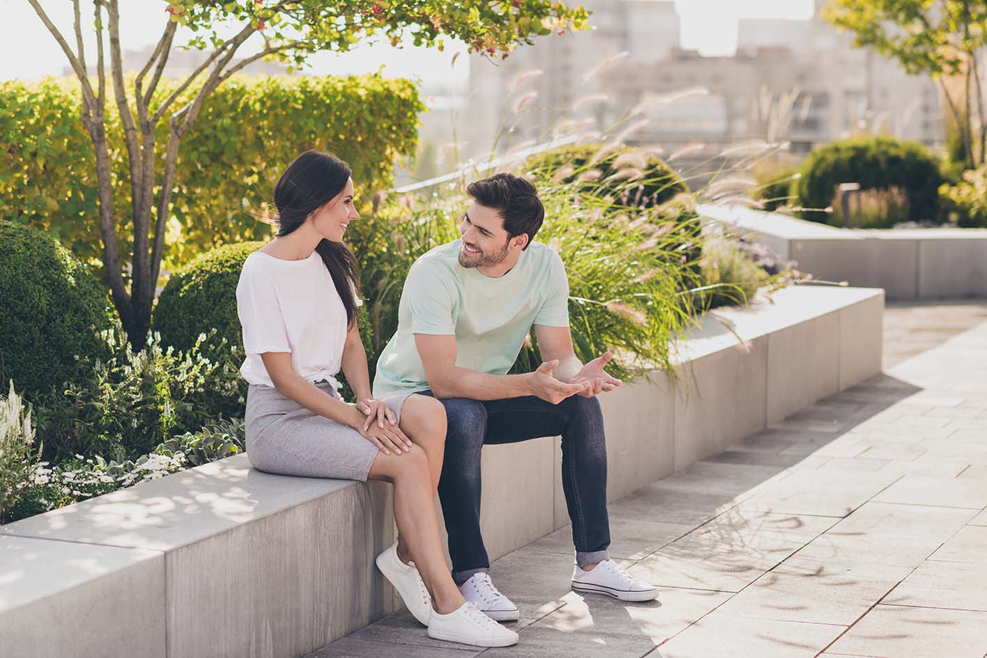 Ζευγάρι καθισμένοι σε πεζούλι με λουλούδια πίσω τους μιλάνε. Στην πόλη