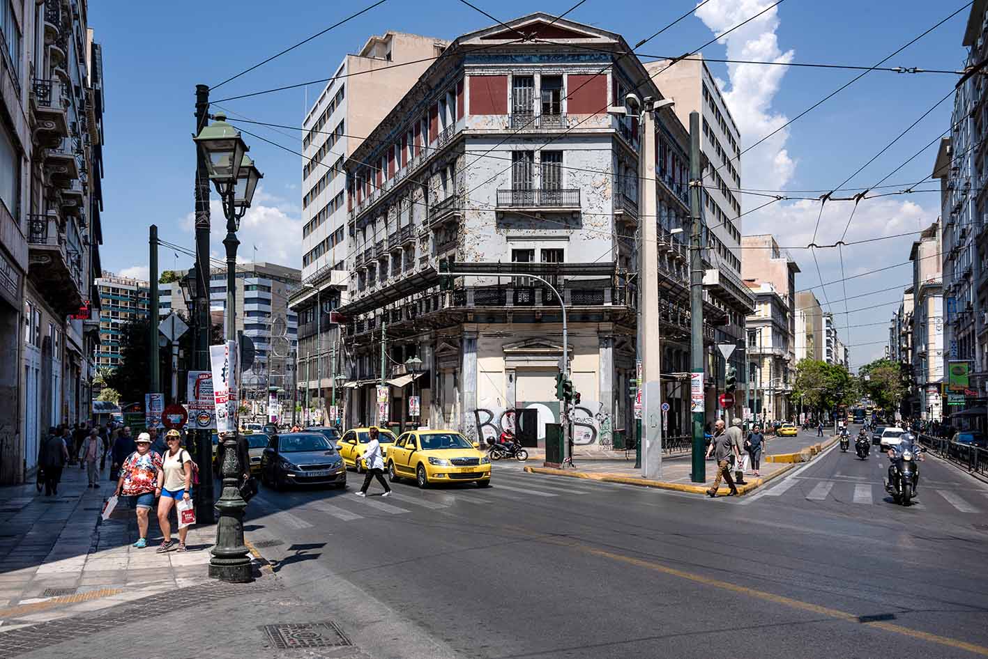 Αθήνα, κοντά στην πλατεία Ομονοίας: Σκηνή δρόμου στο κέντρο της πόλης με αυτοκίνητα, κίτρινα ταξί, κίνηση,  κατοίκους, καταστήματα, κτίρια και γαλάζιο ουρανό στο φόντο. 