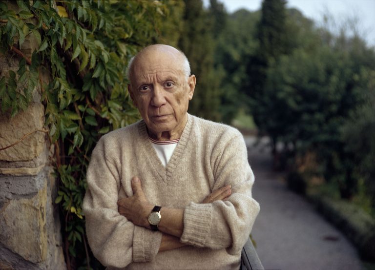 Ο Pablo Picasso στέκεται δίπλα σε μια πράσινη φτέρη με σταυρωμένα χέρια, φορώντας ένα κασμιρένιο πουλόβερ. Φωτογραφία του 1966.