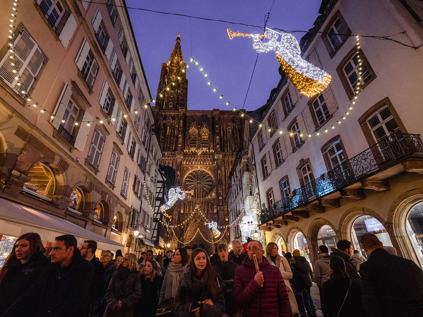 Στρασβούργο, Γαλλία - 24 Δεκεμβρίου 2018: Τουρίστες και ντόπιοι περπατούν στην οδό Merciere με τον καθεδρικό ναό της Notre-Dame de Strasbourg με φόντο χριστουγεννιάτικες φωτισμένες διακοσμήσεις.