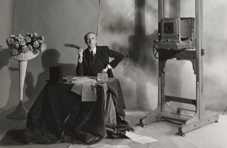 Αυτοπροσωπογραφία του φωτογράφου Cecil Beaton σε τραπέζι με στυλό στο χέρι, σύνθεση λουλουδιών σε ψάθινο καλάθι στα αριστερά και αντίκα φωτογραφική μηχανή στα δεξιά, περίπου τη δεκαετία του 1940.