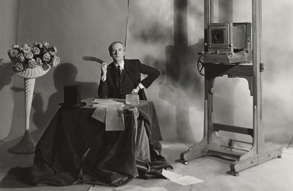 Αυτοπροσωπογραφία του φωτογράφου Cecil Beaton σε τραπέζι με στυλό στο χέρι, σύνθεση λουλουδιών σε ψάθινο καλάθι στα αριστερά και αντίκα φωτογραφική μηχανή στα δεξιά, περίπου τη δεκαετία του 1940.