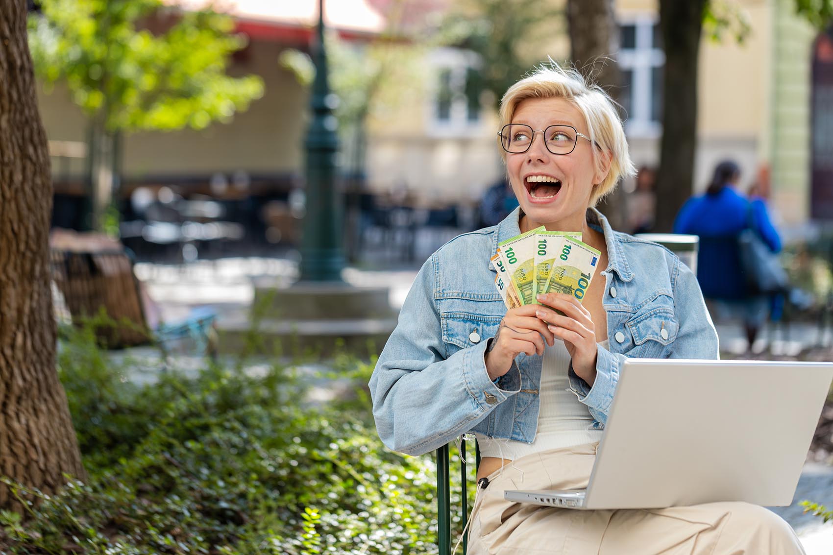 Γυναίκα σε καφέ με κομπιούτερ κρατά αρκετά ευρώ στο χέρι της και γελά χαρούμενα