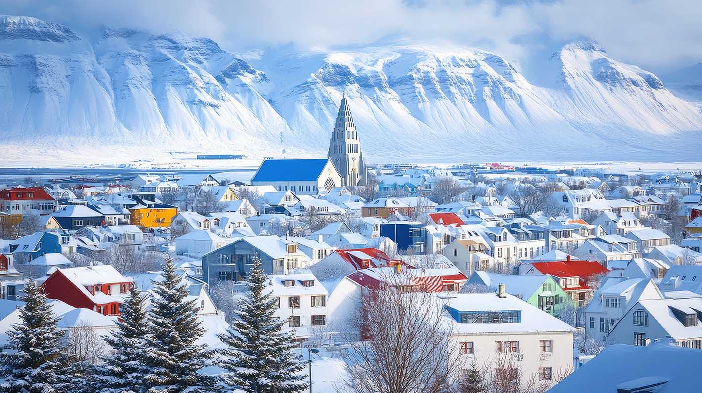 Πανοραμική θέα του Ρέικιαβικ κάτω από ένα στρώμα χιονιού, με πολύχρωμες στέγες και την εμβληματική εκκλησία Hallgrimskirkja να υψώνεται από πάνω. 