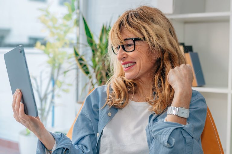 Γυναίκα μέσης ηλικίας παίζει με τα σγουρά μαλλιά της και χαζεύει χαμογελώντας το laptop της