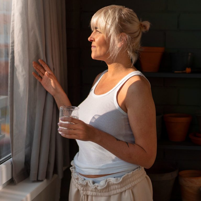 Γυναίκα κοιτά έξω από το παράθυρο κρατώντας ένα ποτήρι νερό