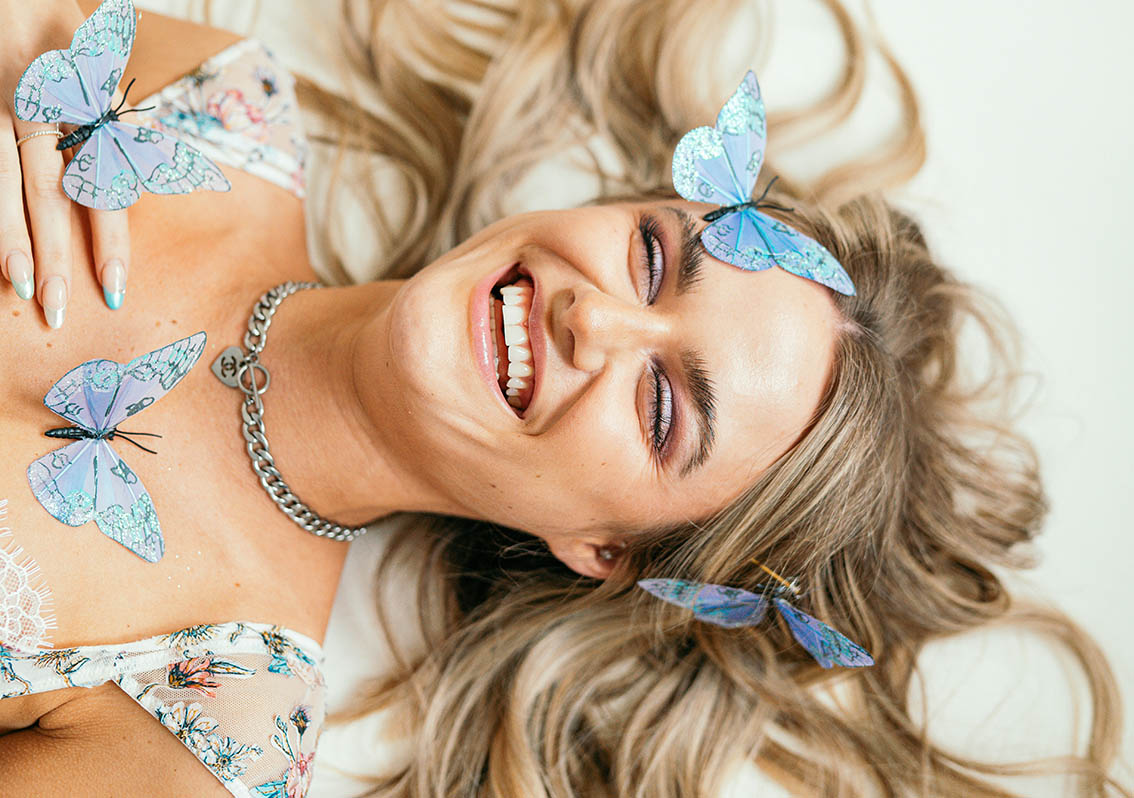 Γυναίκα ξανθιά με ψεύτικες πεταλούδες στο πρόσωπό της χαμογελά ξαπλωμένη στο πάτωμα.