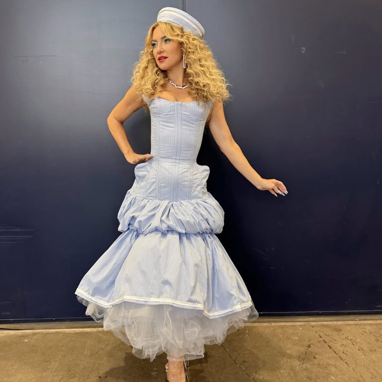 Η Kate Hudson σε εμφάνιση ως κριτής στο RuPaul's Drag Race All Stars. Φοράει ένα γαλάζιο κορσέ-φόρεμα με φουσκωτή φούστα που αποκαλύπτει λευκό τούλι, λευκό ναυτικό καπέλο με ρίγες και έχει έντονα, φουντωτά '70s σγουρά μαλλιά.