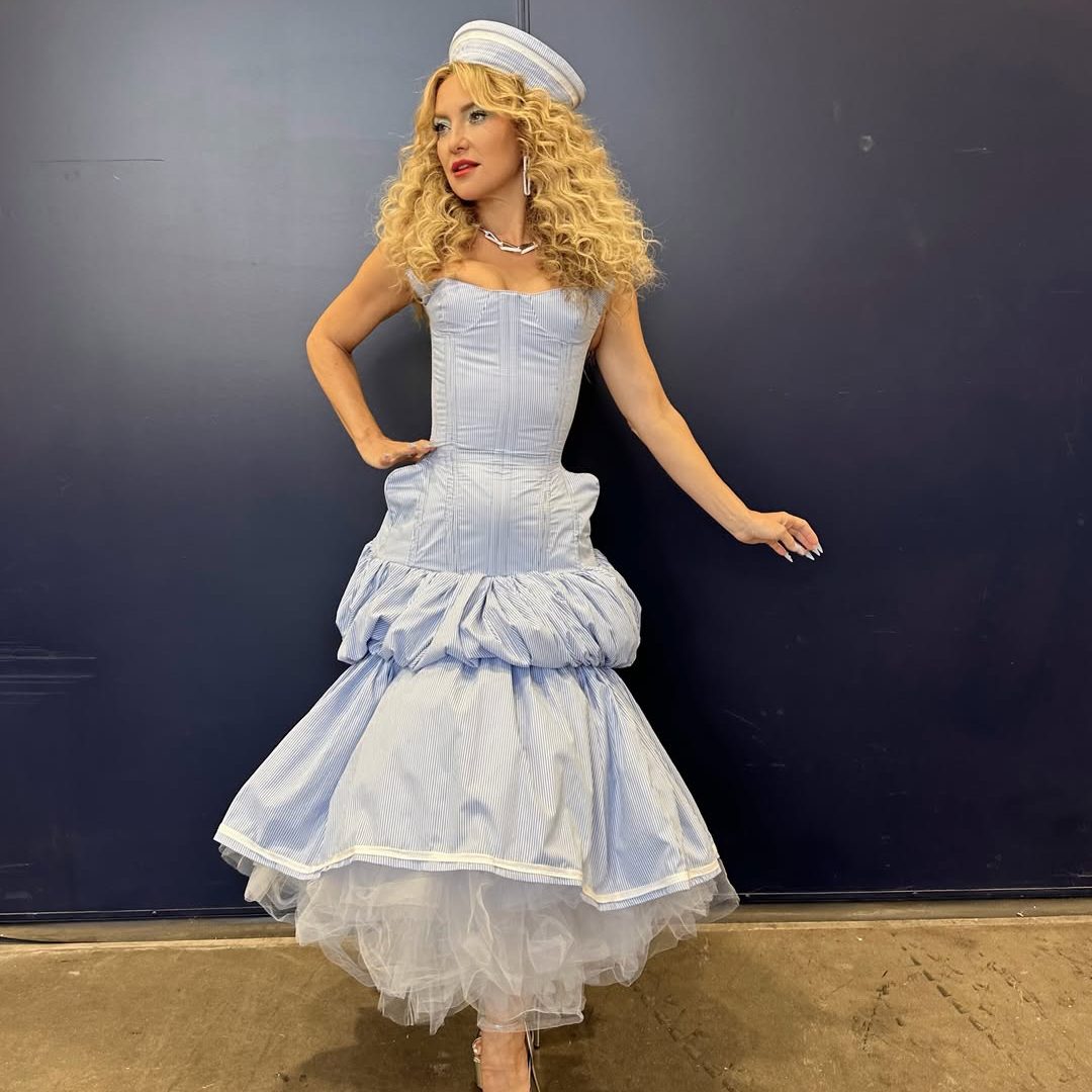 Η Kate Hudson σε εμφάνιση ως κριτής στο RuPaul's Drag Race All Stars. Φοράει ένα γαλάζιο κορσέ-φόρεμα με φουσκωτή φούστα που αποκαλύπτει λευκό τούλι, λευκό ναυτικό καπέλο με ρίγες και έχει έντονα, φουντωτά '70s σγουρά μαλλιά.