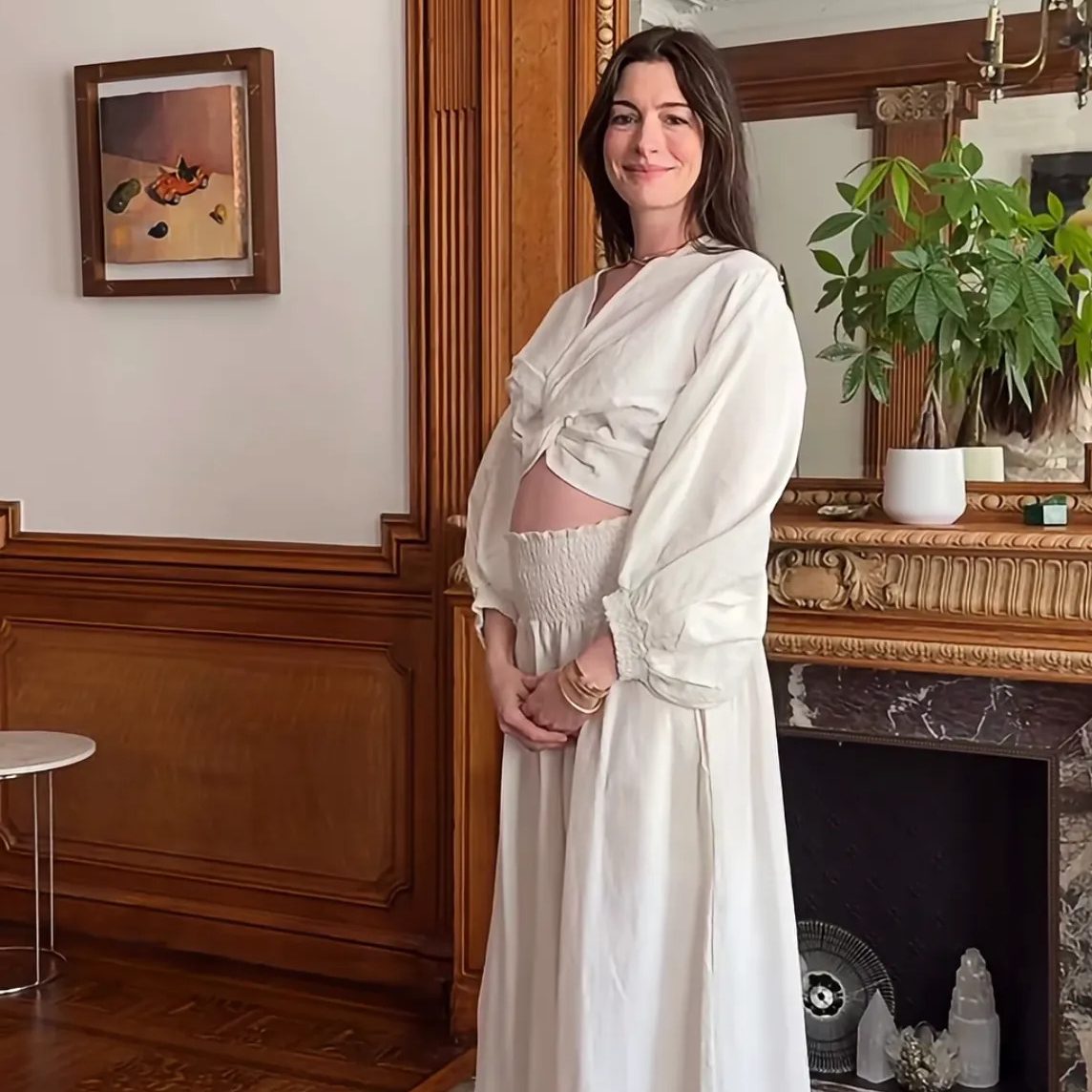 Η Anne Hathaway με λευκή μάξι φούστα, crop τοπ και σανδάλια, να κρατάει τη φουσκωμένη κοιλιά της στην ανακοίνωση της εγκυμοσύνης της στο Instagram.