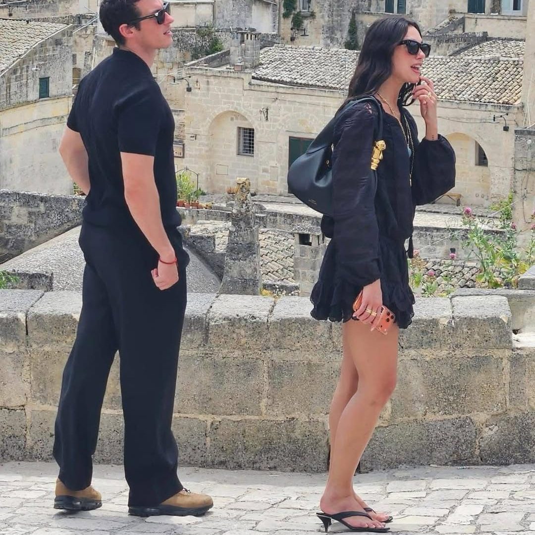 Η Dua Lipa στην πόλη Λούκκα της Ιταλίας με μαύρο μίνι boho φόρεμα, kitten-heel σαγιονάρες και μαύρη δερμάτινη puff τσάντα ώμου με χρυσό χερούλι.