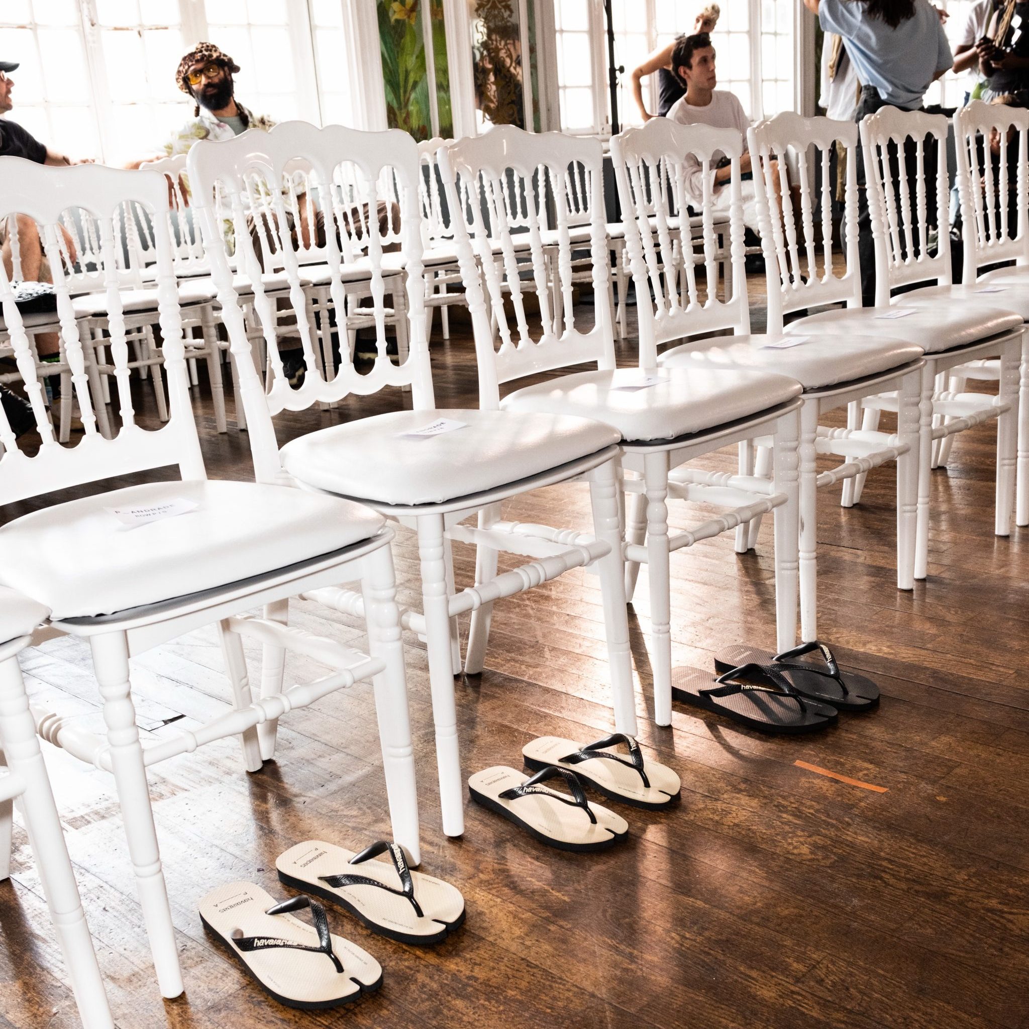 Ζευγάρια σαγιονάρες Havaianas τοποθετημένα κάτω από λευκές καρέκλες στην πρώτη σειρά επίδειξης μόδας στο Παρίσι.