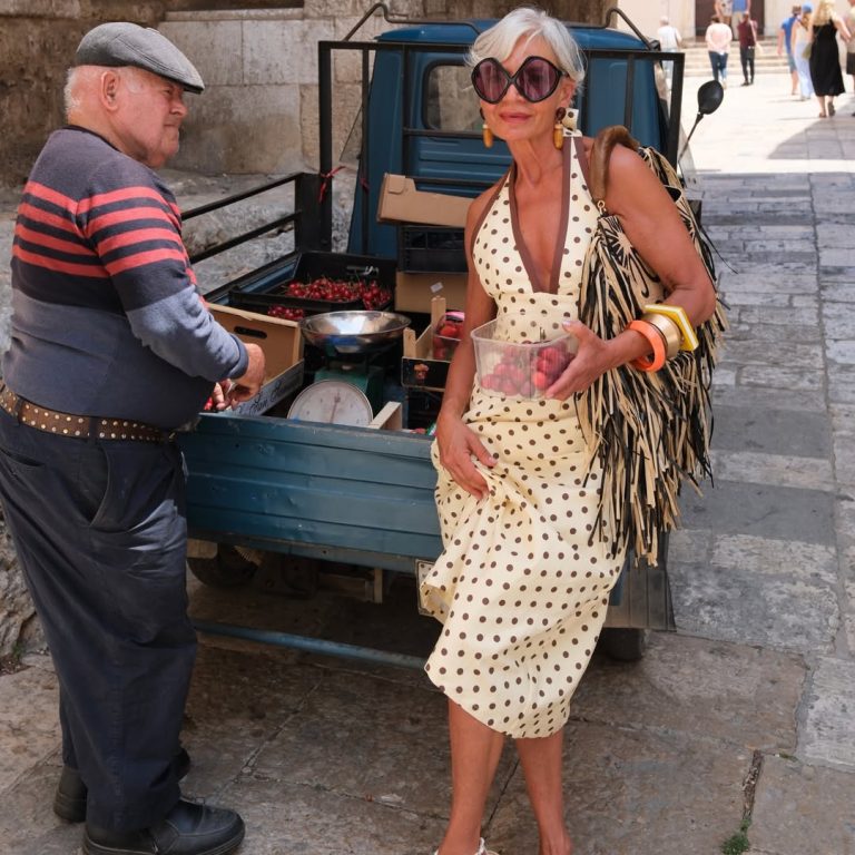 H Grece Ghanem με πουά μίντι φόρεμα σε μπεζ απόχρωση, τσάντα με ψάθινα κρόσσια και bug-eye γυαλιά ηλίου.