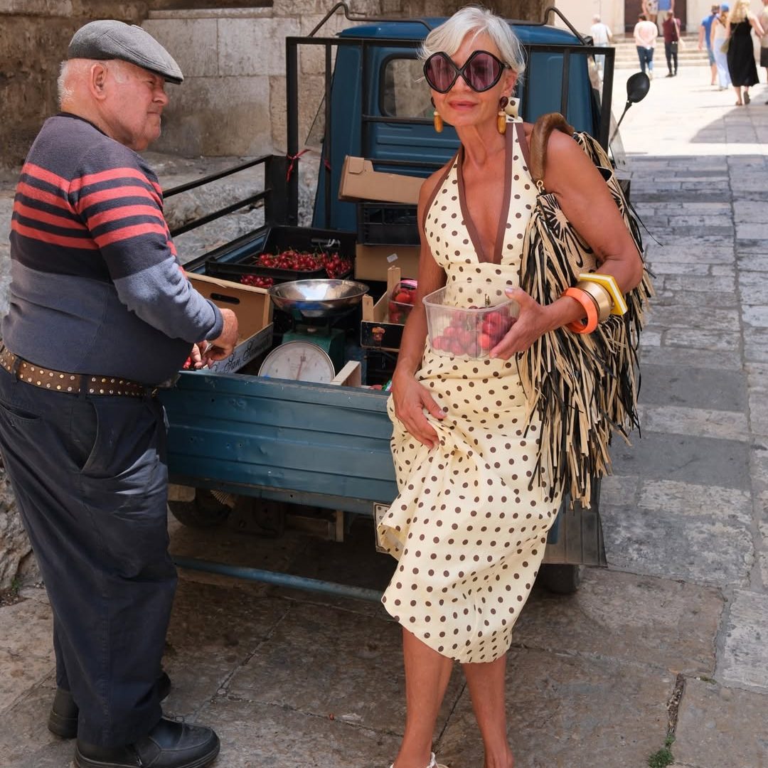 H Grece Ghanem με πουά μίντι φόρεμα σε μπεζ απόχρωση, τσάντα με ψάθινα κρόσσια και bug-eye γυαλιά ηλίου.