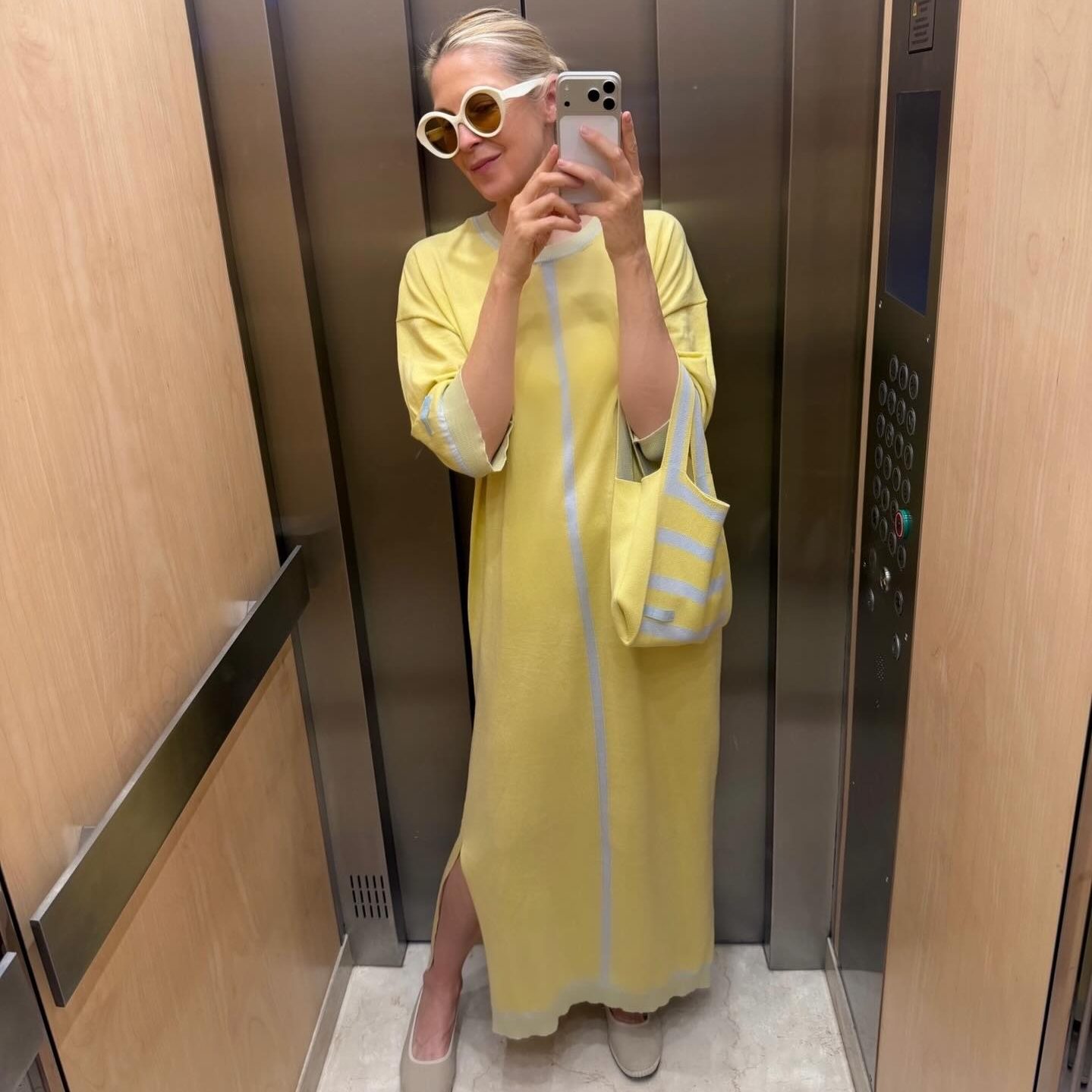 Μια selfie της Kelly Rutherford μέσα σε ασανσέρ, να φοράει ένα μακρύ, πλεκτό κίτρινο φόρεμα με γαλάζιες λεπτομέρειες, ασορτί κίτρινη τσάντα Charlotte Beaude, μπεζ μπαλαρίνες Cayumas, λευκά γυαλιά ηλίου και να βγάζει φωτογραφία με το κινητό της.