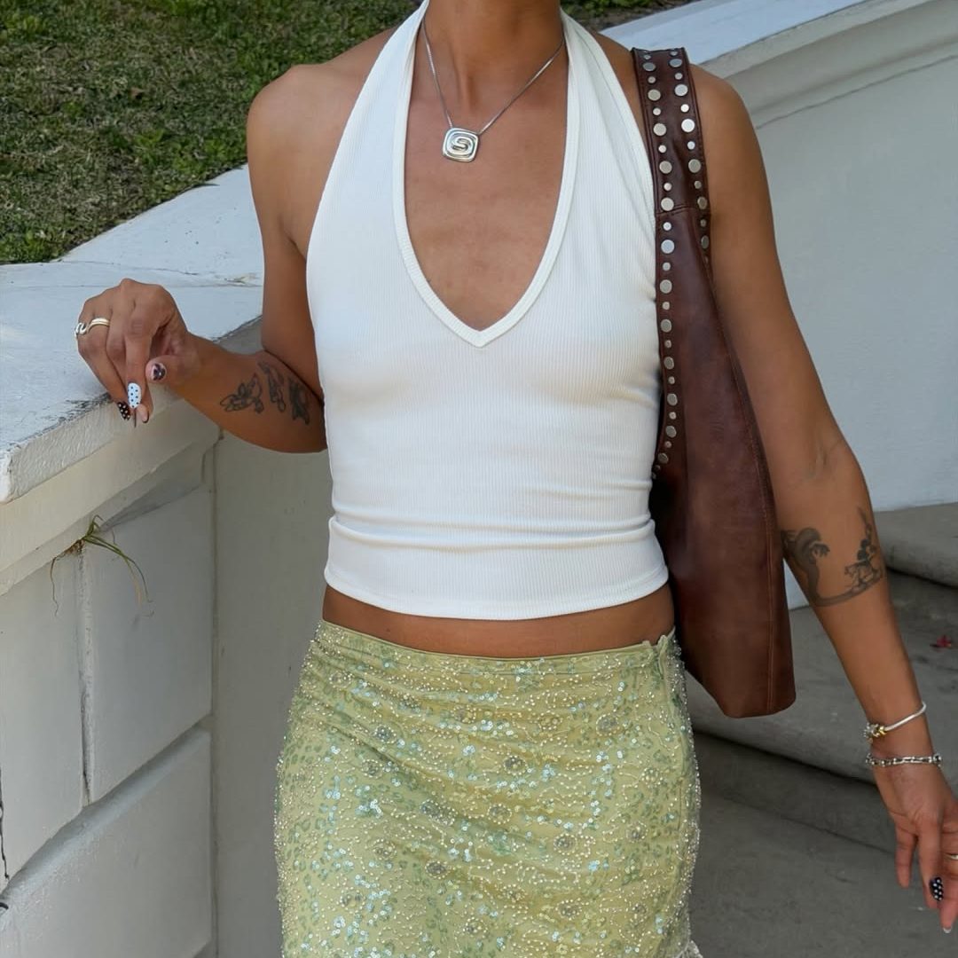 Γυναίκα σε καλοκαιρινό look με λευκό V-neck tank τοπ, μίνι φούστα σε lime απόχρωση με παγιέτες, χάντρες και κρόσσια και καφέ δερμάτινη τσάντα ώμου με τρούκς στο λουράκι της.