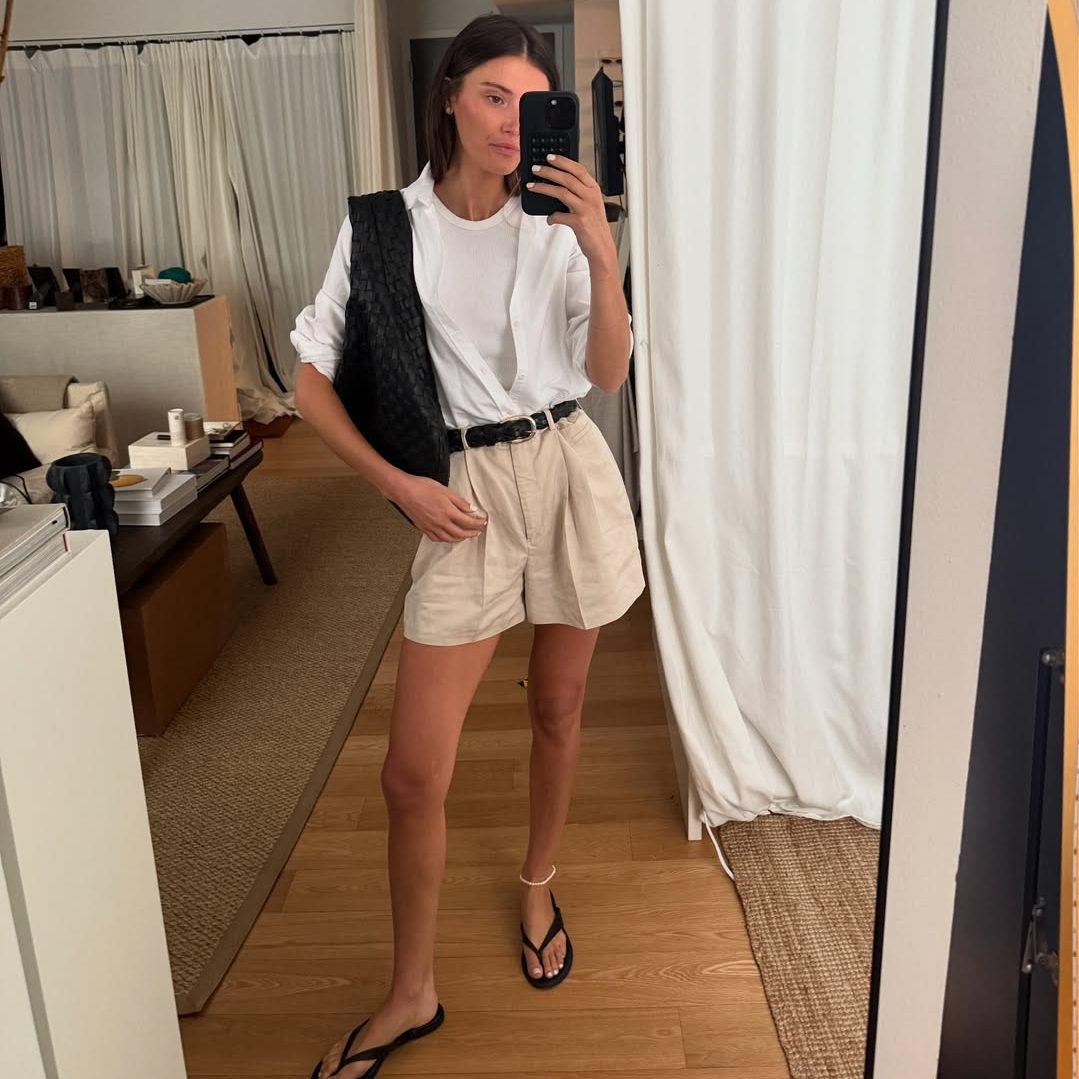 Street style έμπνευση με λευκό tank τοπ, ανοιχτό λευκό πουκάμισο και ψηλόμεσο μπεζ λινό σορτς με μαύρη ζώνη, μαύρες σαγιονάρες και oversized μαύρη τσάντα ώμου.