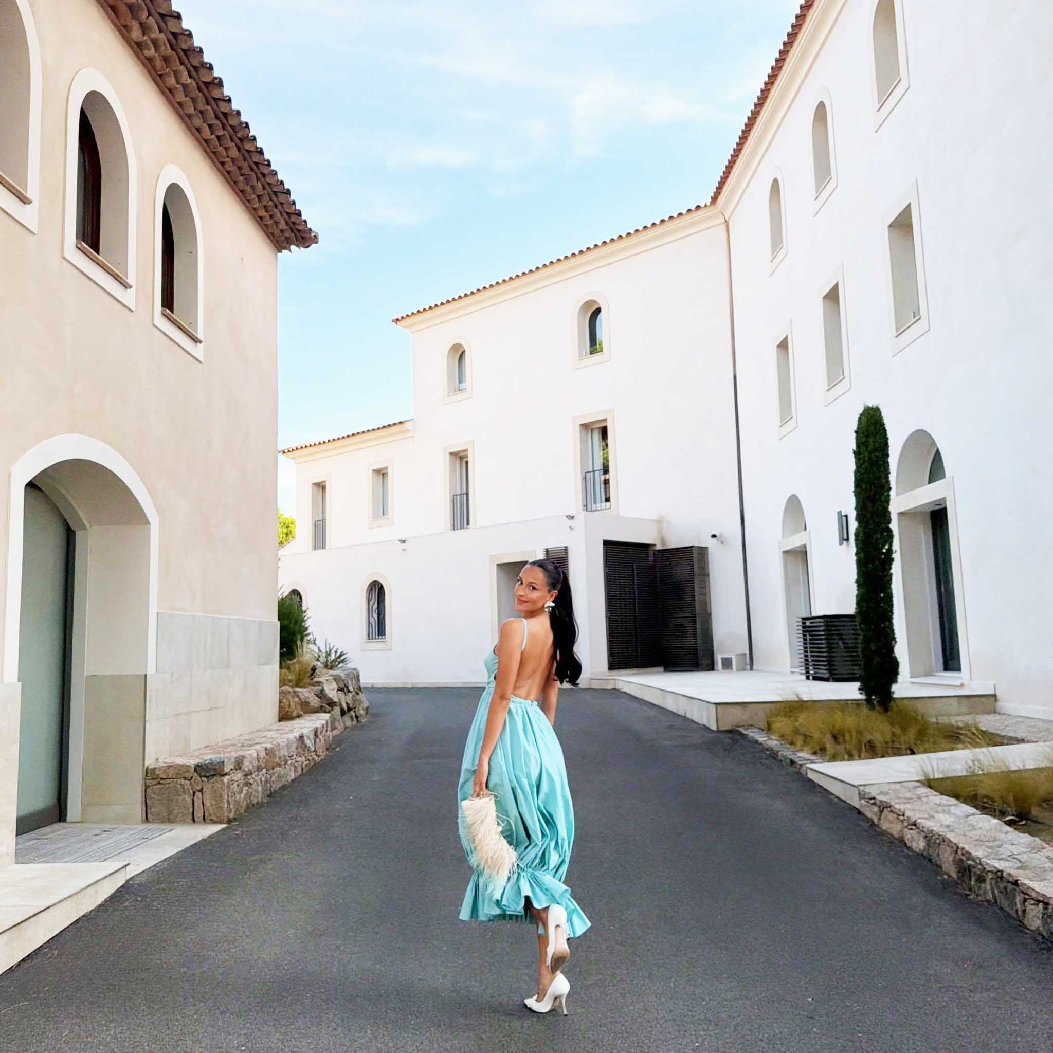 Η Alexandra Leclerc σε εμφάνιση με εξώπλατο μπαλούν φόρεμα σε απόχρωση aqua blue, κρατώντας λευκή τσάντα με φτερά.