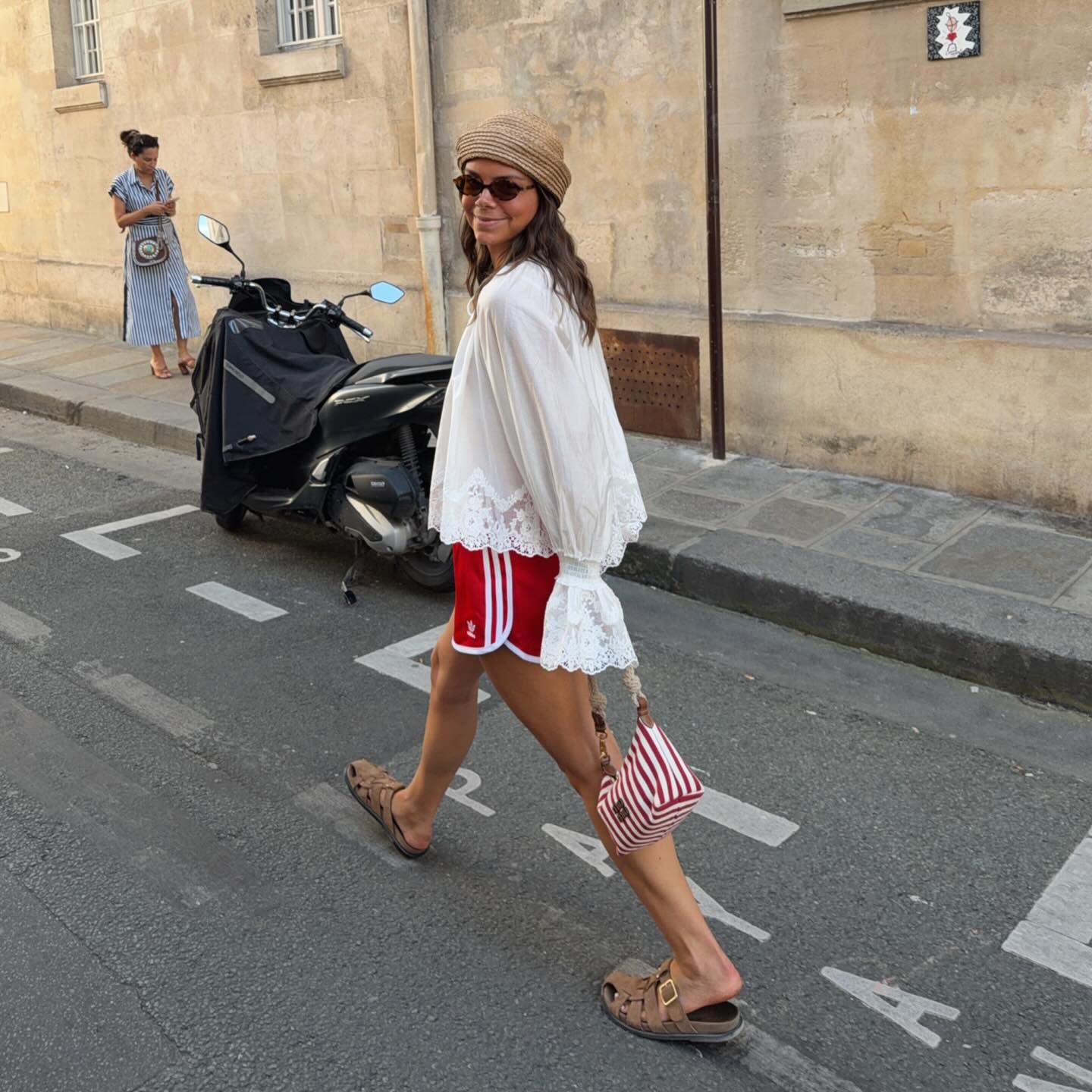 Γυναίκα με κόκκινο αθλητικό σορτς adidas, λευκή boho μπλούζα με δαντέλα, ψάθινο καπέλο και ριγέ τσάντα.