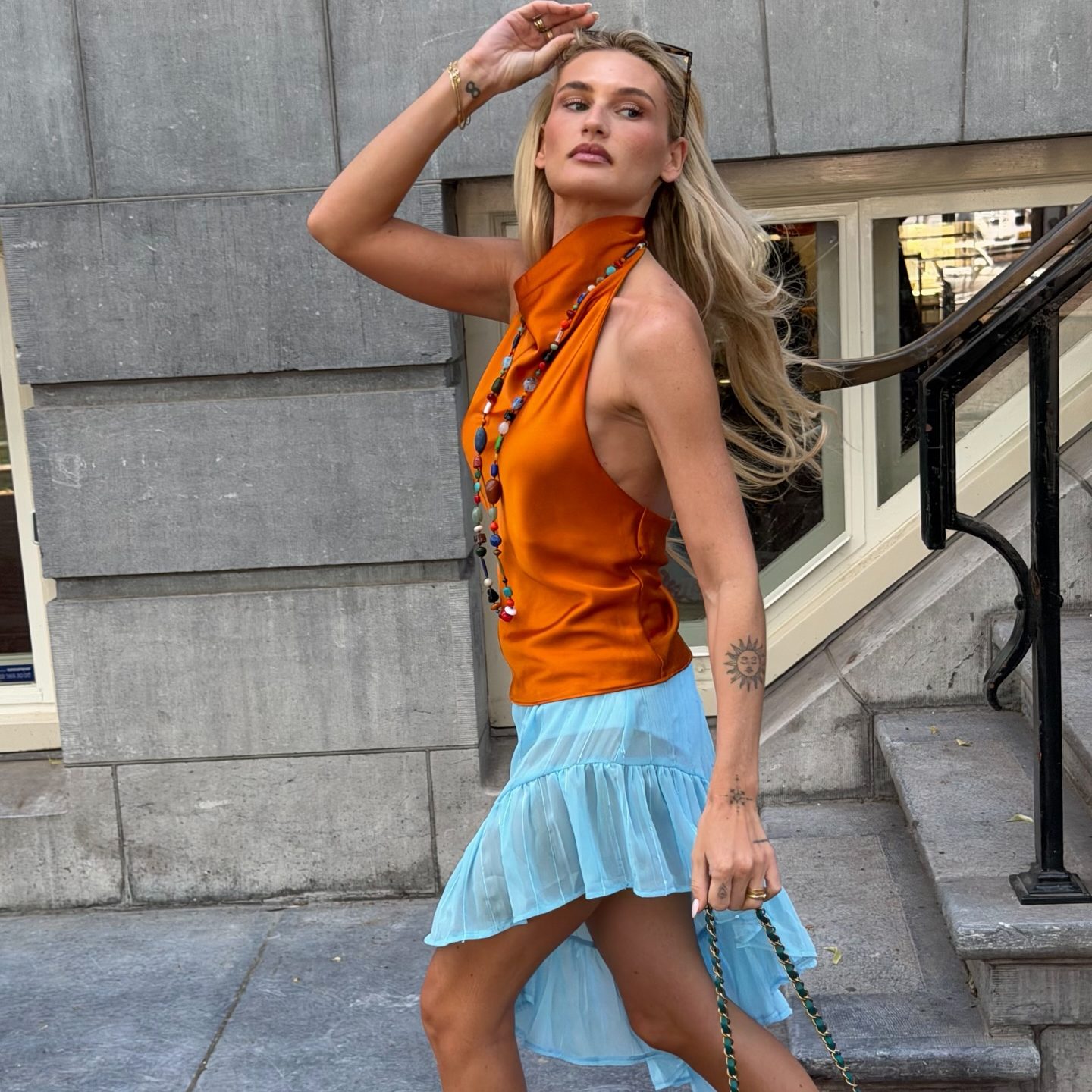 Γυναίκα με colormaxxing look που φοράει πορτοκαλί σατέν τοπ, γαλάζια ασύμμετρη φούστα, κερασί πέδιλα με τακούνι, πετρόλ τσάντα Chanel και χρωματιστό κολιέ.