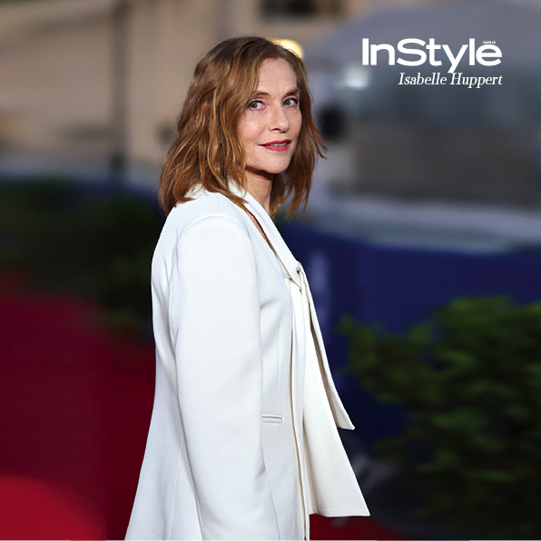 η Isabelle Huppert σε κόκκινο χαλί, ντυμένη με λευκό κοστούμι, κοιτάζει προς την κάμερα με φόντο αστικό περιβάλλον