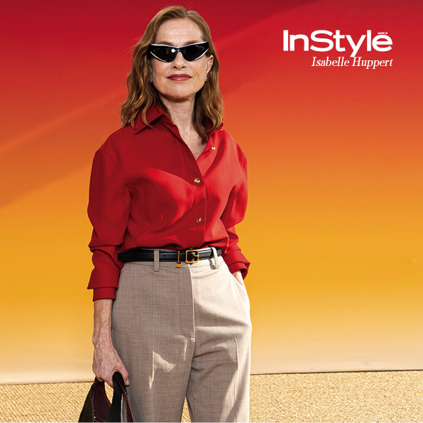 η Isabelle Huppert με κόκκινο πουκάμισο, μπεζ παντελόνι και μαύρα γυαλιά ηλίου ποζάρει μπροστά σε έντονο πορτοκαλί-κόκκινο φόντο