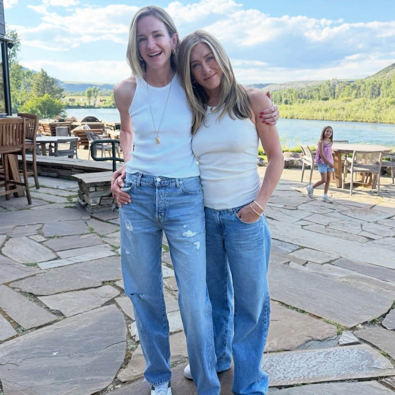 Η Jennifer Aniston με λευκό ριμπ tank τοπ και φαρδύ τζιν παντελόνι αγκαλιάζει μια φίλη της με παρόμοιο look σε εξωτερικό χώρο.