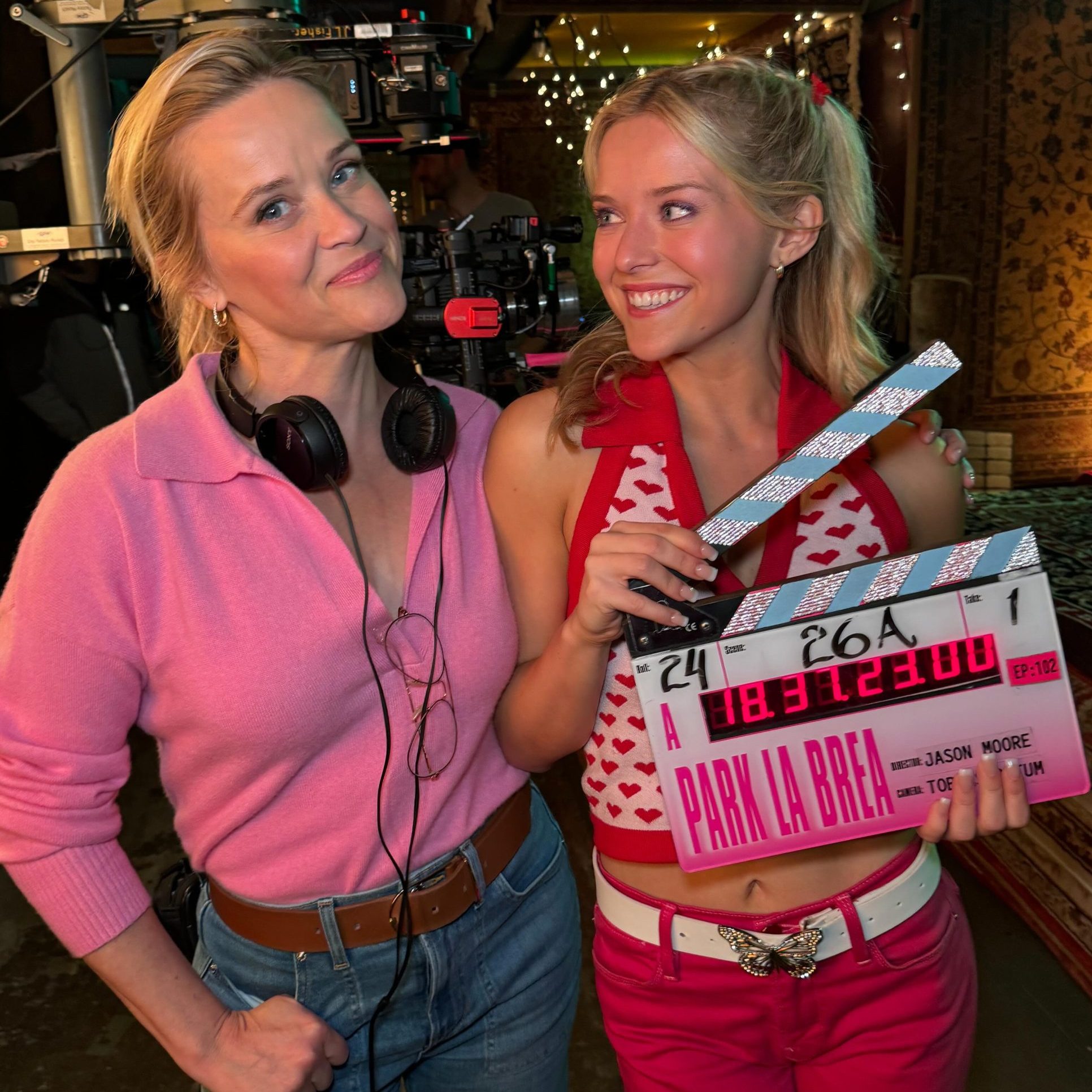 Η Reese Witherspoon με ροζ πουλόβερ και τζιν στέκεται δίπλα στη Lexi Minetree, η οποία κρατάει μια ροζ κλακέτα της σειράς "Park La Brea" στα παρασκήνια των γυρισμάτων.