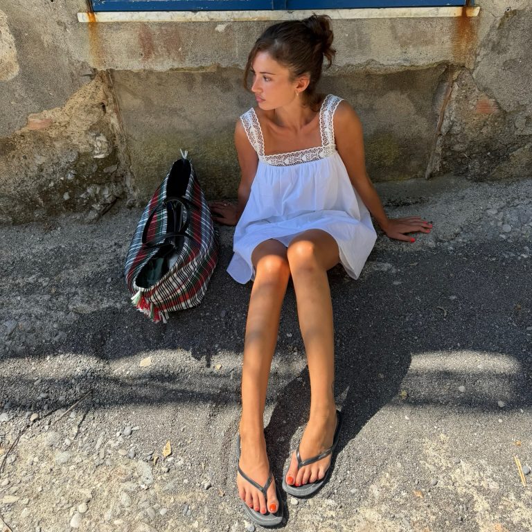 Μια κοπέλα φοράει ένα κοντό λευκό φόρεμα με δαντέλα, μαύρες σαγιονάρες και έχει δίπλα της μια καρό υφαντή τσάντα.