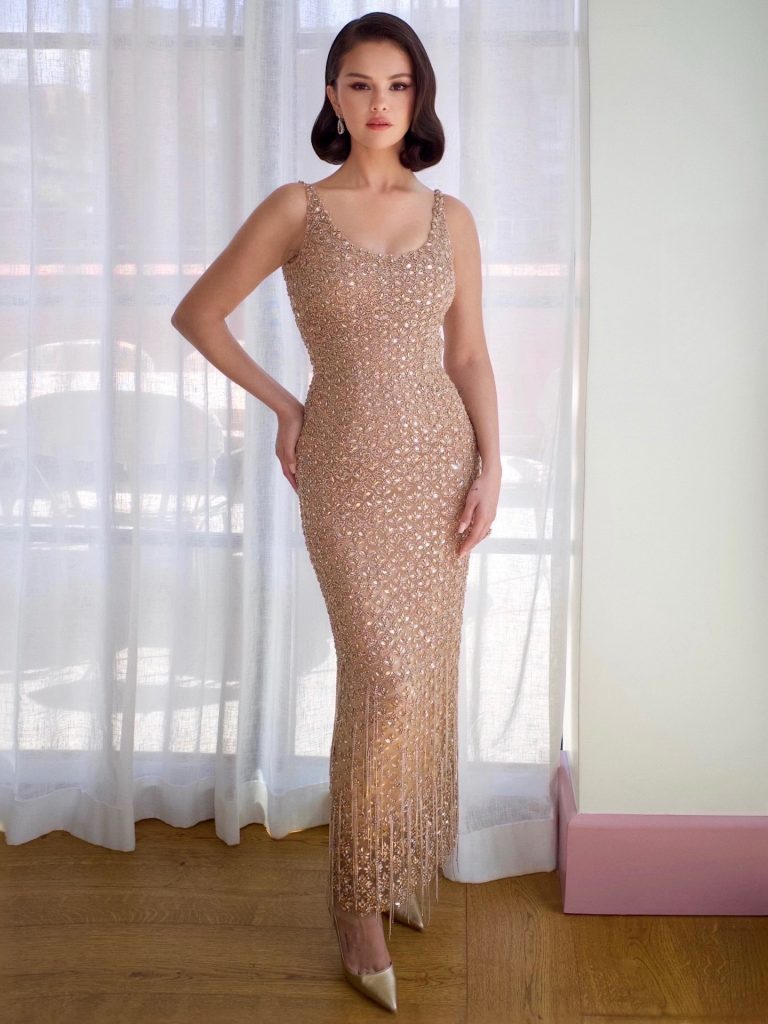 Η Selena Gomez ποζάρει μπροστά από ένα φωτεινό παράθυρο, με τη στενή, μάξι δημιουργία Oscar de la Renta, η οποία είναι καλυμμένη με λαμπερούς κρυστάλλους και διαθέτει ένα χαρακτηριστικό τελείωμα με χρυσά κρόσσια.