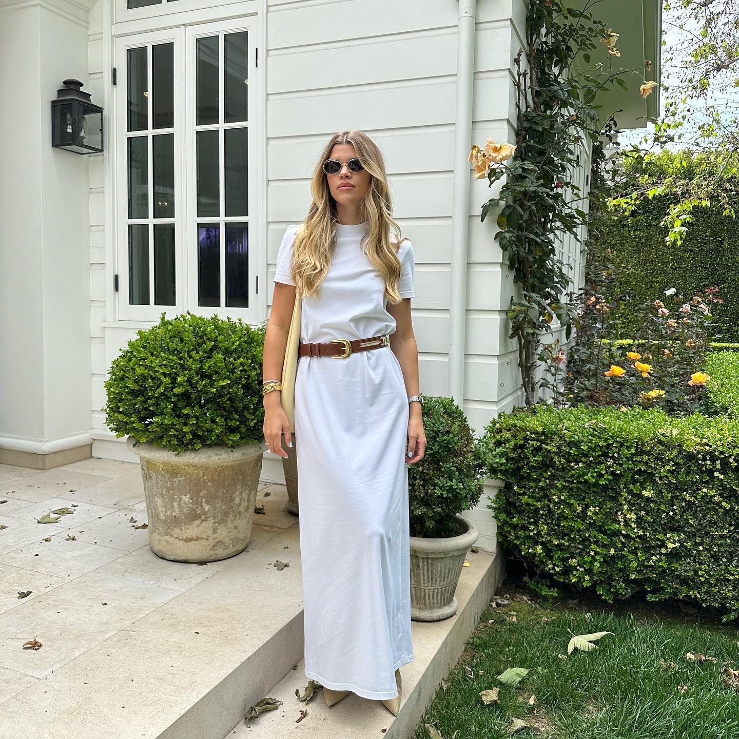 Η Sofia Richie Grainge με μάξι λευκό t-shirt φόρεμα, καφέ δερμάτινη ζώνη με χρυσή αγκράφα και μαύρα γυαλιά ηλίου.