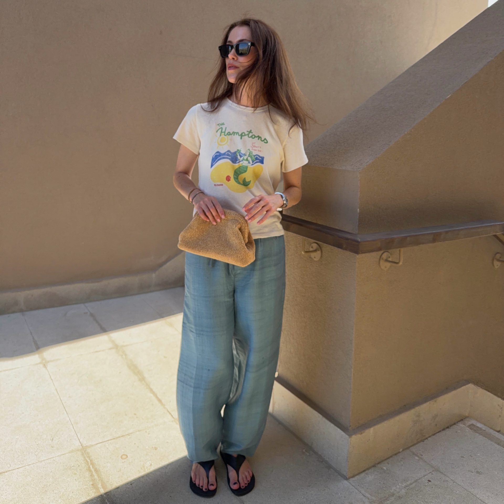 Το λευκό graphic t-shirt συνδυάζεται με λινό φαρδύ παντελόνι και ψάθινο clutch για ένα δροσερό, effortless καλοκαιρινό σύνολο.