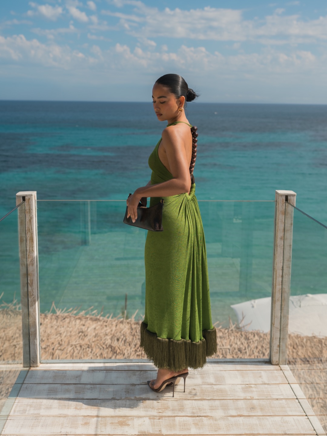 Μια εντυπωσιακή εμφάνιση με λαδί backless μάξι φόρεμα με κρόσσια στο τελείωμα και ξύλινες χάντρες στην πλάτη, ιδανικό για καλοκαιρινό destination γάμο με φόντο το απέραντο γαλάζιο.