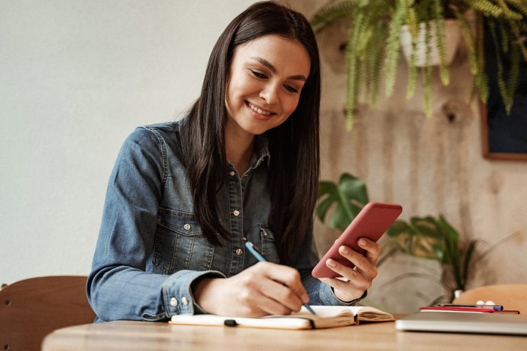Χαρούμενη γυναίκα που κρατά smartphone και γράφει σε σημειωματάριο