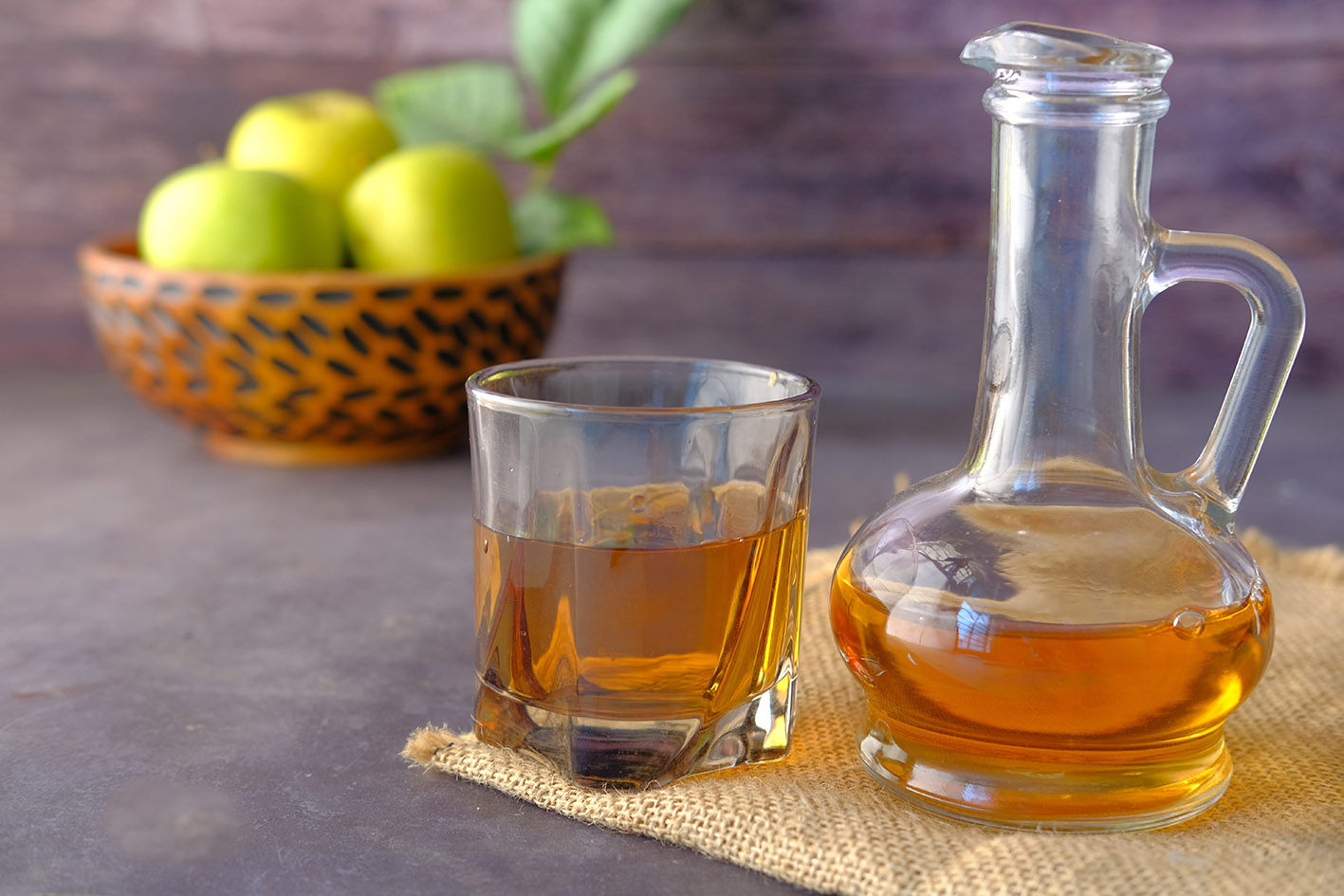 Μηλόξιδο σε ποτήρι και μπουκάλι. Στο βάθος της φωτογραφίας ένα καλάθι με μήλα πράσινα.