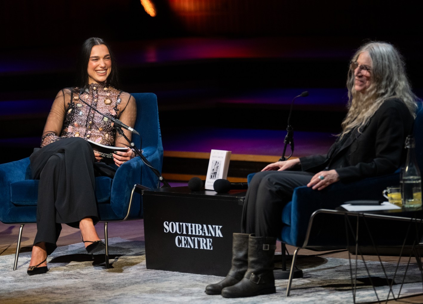 Η Dua Lipa και η Patti Smith εμφανίζονται στη σκηνή κατά τη διάρκεια της εκδήλωσης «Dua Lipa In Conversation With Patti Smith» στο Φεστιβάλ Λογοτεχνίας του Λονδίνου 2026, στο Royal Festival Hall, στις 25 Οκτωβρίου 2026, στο Λονδίνο Για την εκδήλωση επέλεξε διάφανο κεντημένο τοπ Schiaparelli, μαύρο παντελόνι και χρυσή αγκράφα σε σχήμα χταποδιού.