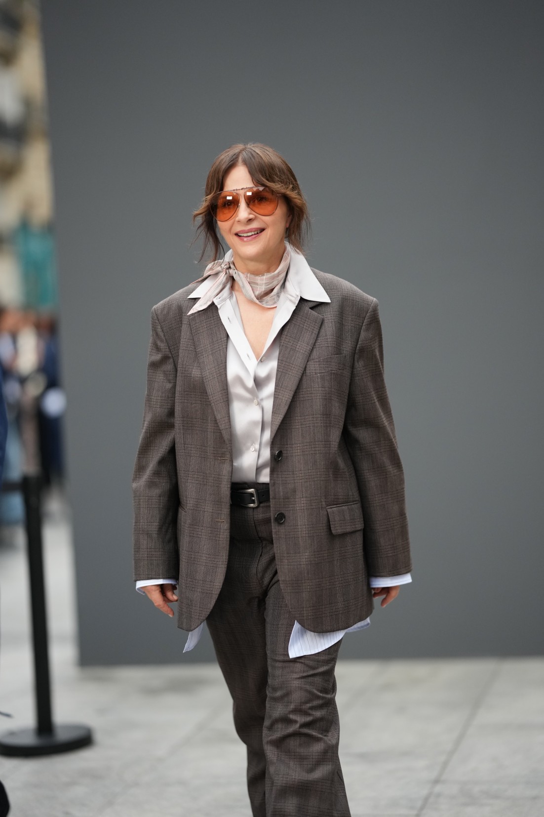 Η Juliette Binoche εμφανίζεται με κοντά, κυματιστά καστανά μαλλιά, πορτοκαλί γυαλιά ηλίου τύπου «aviator» με καφέ φακούς, ένα ροζ φουλάρι με διακριτικό καρό μοτίβο και ένα καφέ καρό σακάκι. Από μέσα φορά λευκό μεταξωτό πουκάμισο με κουμπιά. Η φωτογραφία τραβήχτηκε έξω από το κατάστημα Acne Studios, κατά τη διάρκεια της Εβδομάδας Μόδας του Παρισιού για την Άνοιξη/Καλοκαίρι 2027, στις 30 Σεπτεμβρίου 2026.