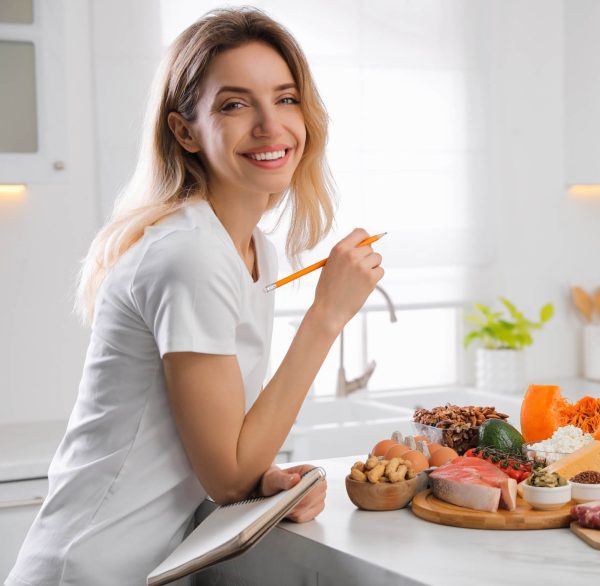 Woman with notebook and healthy food at white table in kitchen. Keto diet