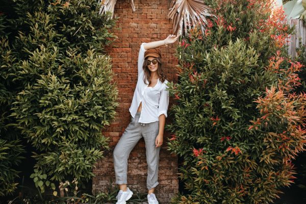 Slim lady in white sneakers, gray pants and white oversized blouse poses against brick wall surroun