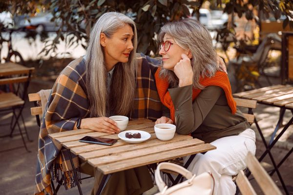 Empathic grey haired Asian woman hugs friend to cheer up at table in street cafe
