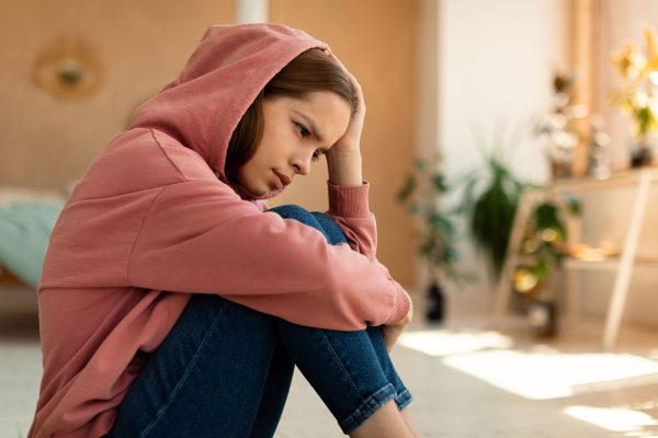 Portrait of upset teen girl feeling upset and depressed, looking away and wearing hood, sitting on floor at home