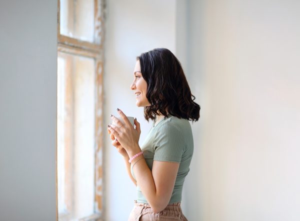 Young happy woman standing indoor with cup of tea looking out window
