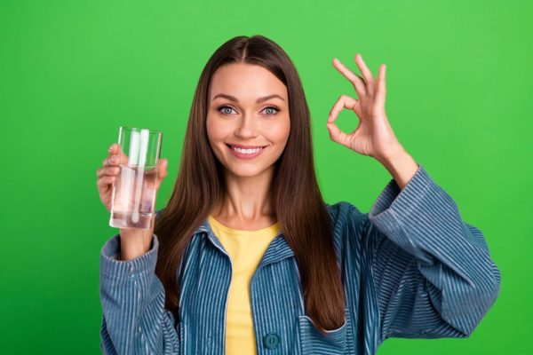 Photo of attractive cheerful lady hold water glass hand fingers demonstrate okey symbol isolated on green color background